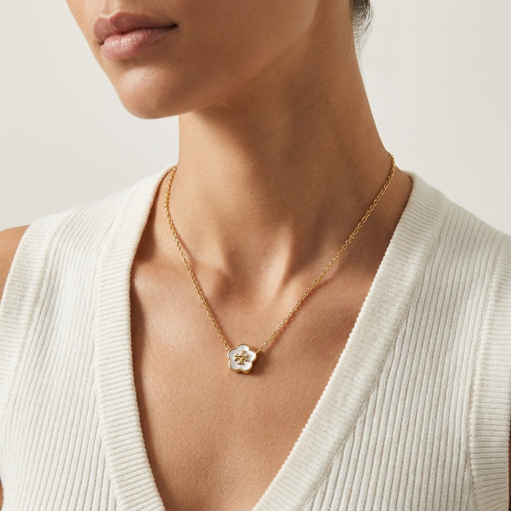 A close-up of a model wearing a delicate gold chain necklace with a small white flower-shaped pendant featuring a gold center, styled with a white ribbed knit top.