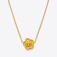 A gold chain necklace featuring a small five-petal flower pendant with a yellow enamel center and a gold monogram logo, shown on a light gray background.