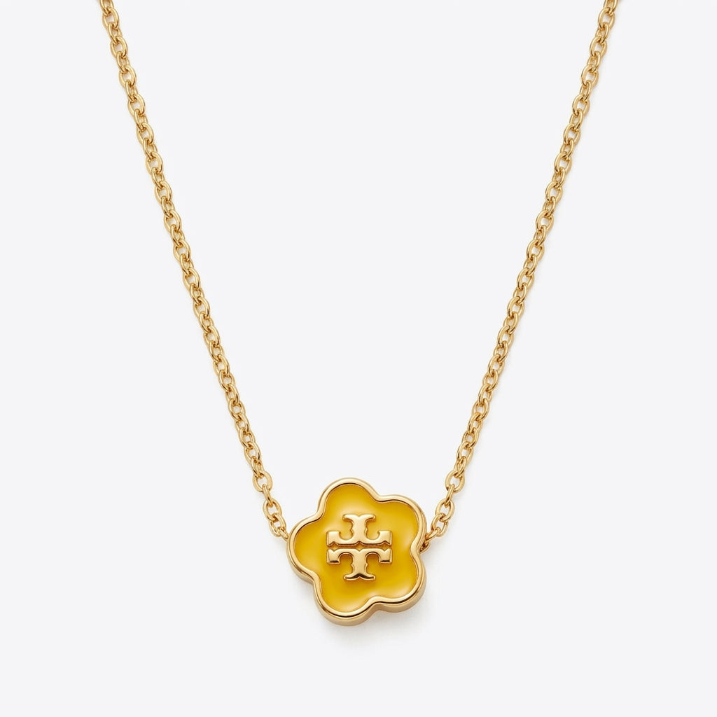A gold chain necklace featuring a small five-petal flower pendant with a yellow enamel center and a gold monogram logo, shown on a light gray background.