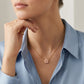 A close-up lifestyle shot of a model wearing a delicate gold chain necklace with a small pink flower pendant, styled with a light blue button-down shirt.