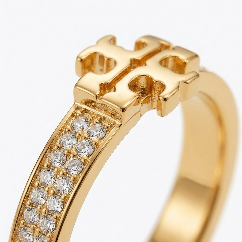 A gold ring featuring a geometric cross-like top and a curved band set with clear pavé stones, shown on a clean white background.