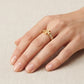 A gold ring with a geometric openwork design and a pavé-set row of small clear stones, shown worn on a hand against a neutral beige textured background.