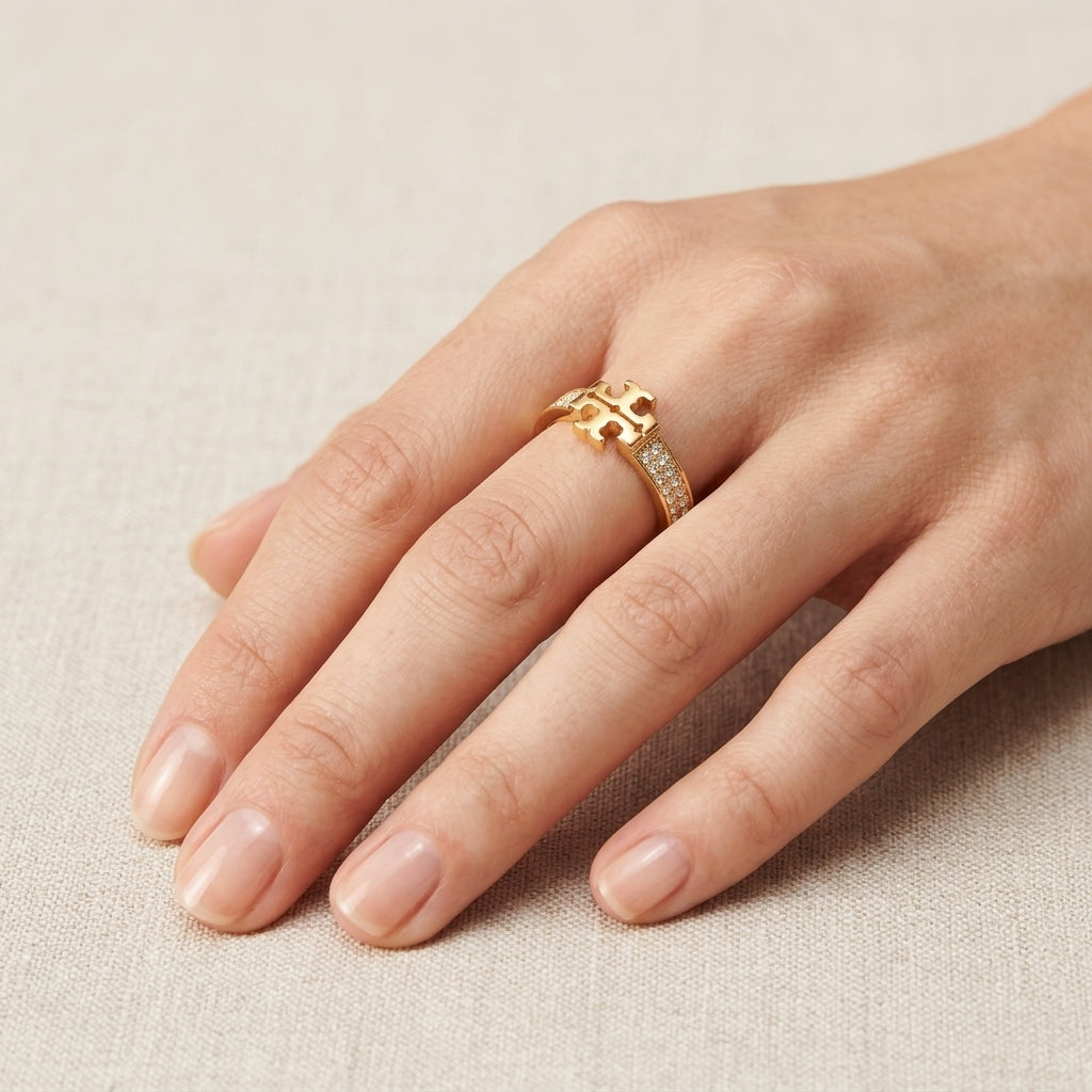 A gold ring with a geometric openwork design and a pavé-set row of small clear stones, shown worn on a hand against a neutral beige textured background.