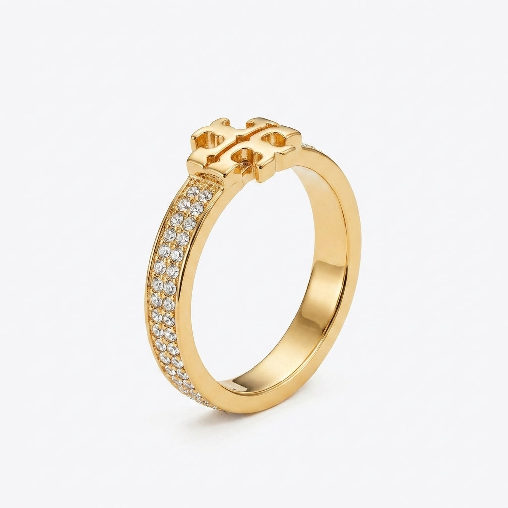 A gold ring featuring a decorative interlocking monogram design at the top and a channel-set row of clear stones along one side, shown on a light gray background.