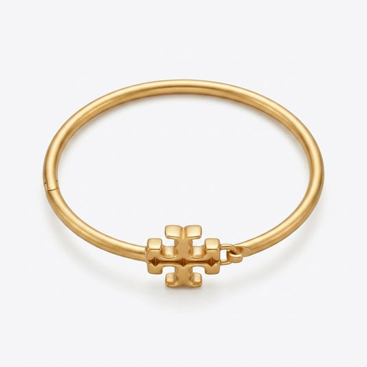 A gold bangle bracelet with a small geometric logo charm, displayed on a clean white background.