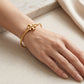 A gold bangle bracelet with a sculptural, openwork floral or abstract flower charm, shown worn on a person's wrist against a neutral beige fabric background.