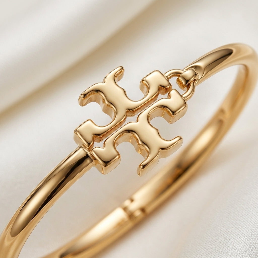 A close-up of a polished gold bangle bracelet featuring a decorative interlocking monogram-style centerpiece, resting on a soft white fabric background.