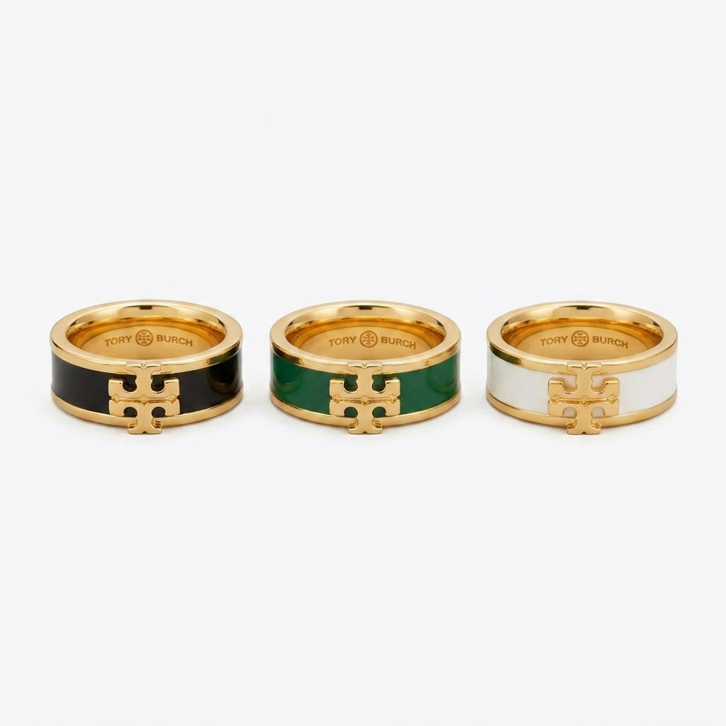 Three Tory Burch rings with gold-tone metal and enamel inserts in black, green, and white, arranged side by side on a light gray background.