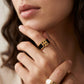 A close-up of a model's hand wearing a gold ring with a black enamel band and a stylized gold logo motif, with a second gold ring partially visible on another finger.
