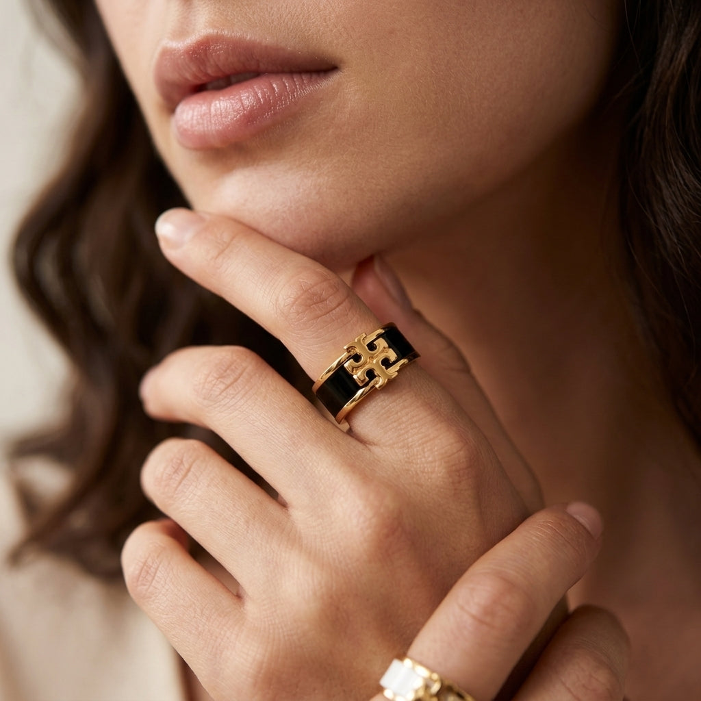 A close-up of a model's hand wearing a gold ring with a black enamel band and a stylized gold logo motif, with a second gold ring partially visible on another finger.