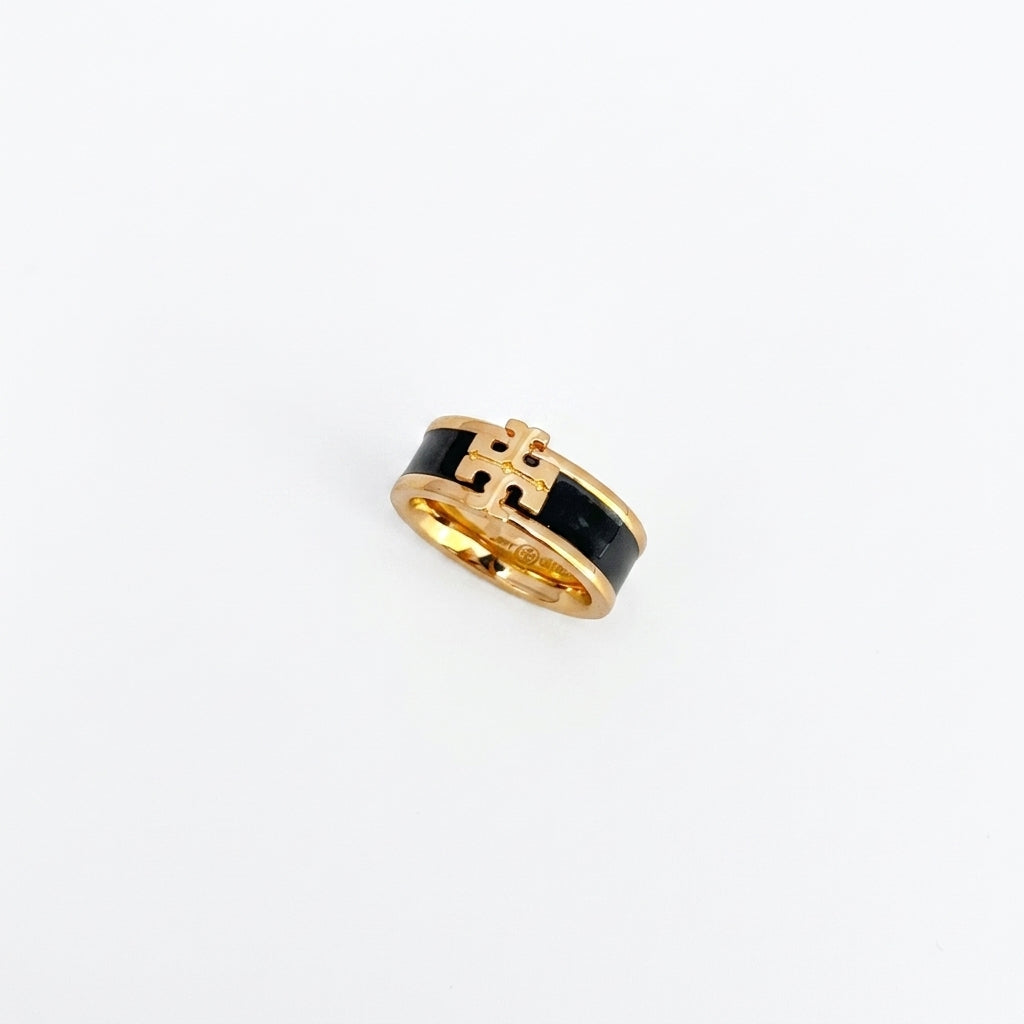 A gold ring with a black enamel inlay and a central interlocking monogram design, photographed on a clean white background.
