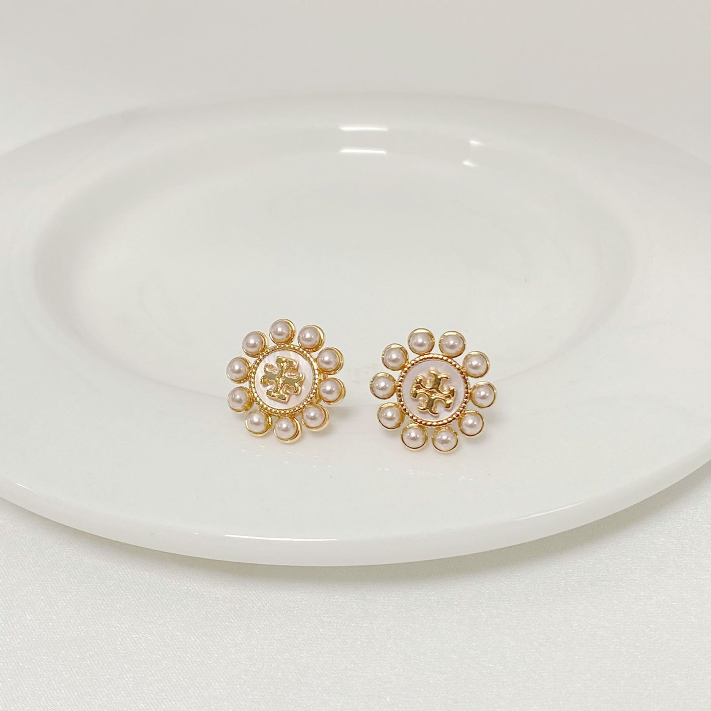 Both studs displayed face-up on a soft, light background (ivory or pale green), highlighting the floral shape and central pearl.
