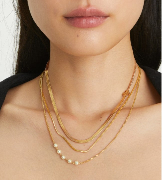 A close-up of a model wearing a layered gold necklace set with three chains, one featuring a row of small white pearls.