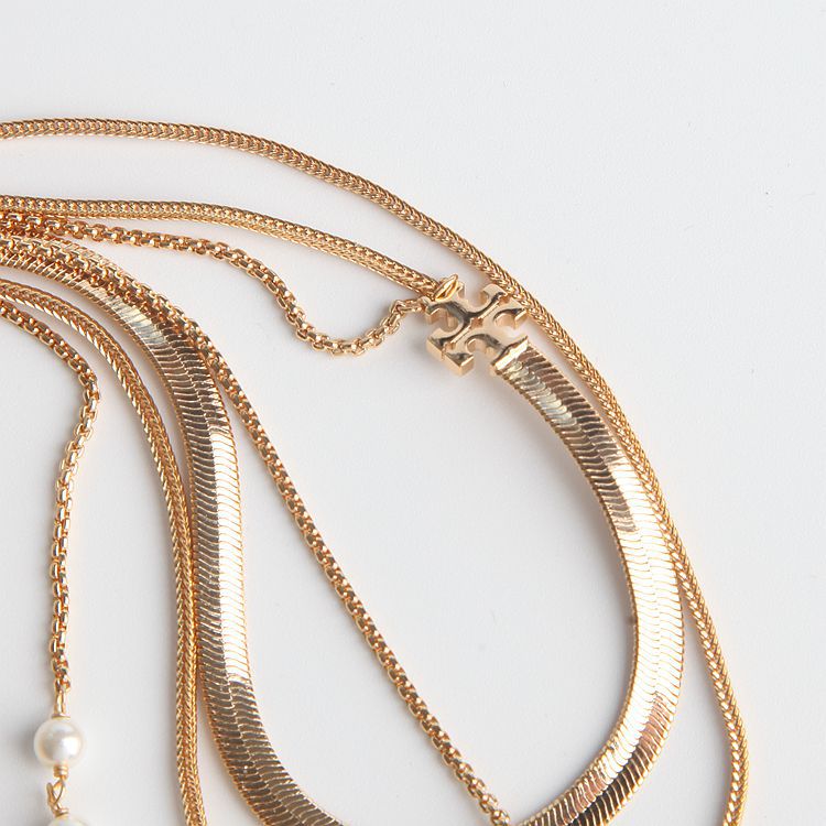 A close-up of layered gold-tone necklaces on a white background, including a snake chain, a textured chain, a delicate chain with a pearl bead, and a small gold H-shaped charm.