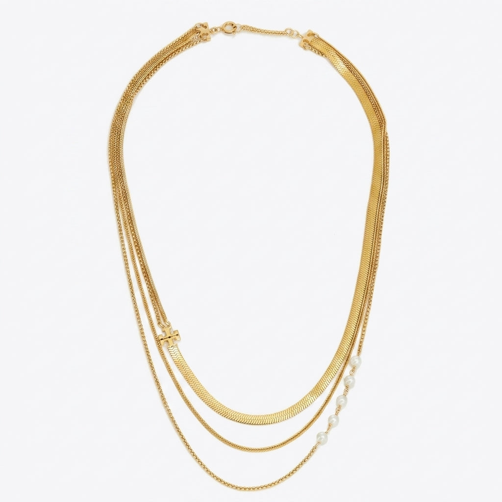 A gold multi-strand necklace featuring three layered chains, including a textured snake chain, a delicate cable chain, and a fine chain accented with small white pearls, shown on a plain white background.