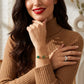 A woman in a brown ribbed turtleneck sweater wearing a delicate gold bracelet with two green circular charms, one with a monogram logo. She is also wearing a silver chevron ring on her finger.