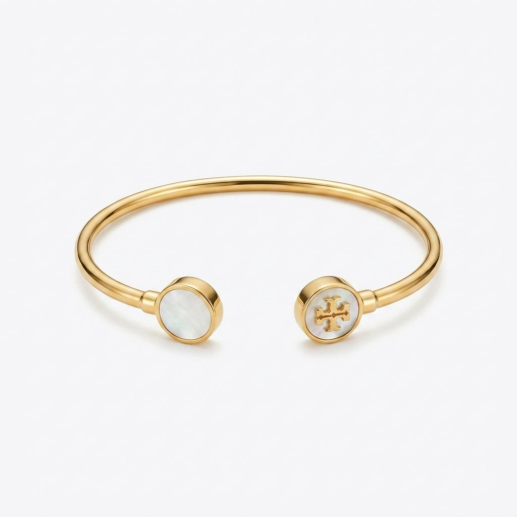 A gold-tone open cuff bangle bracelet with two round bezel-set inlays, one featuring white mother-of-pearl and the other displaying a gold monogram logo, set against a clean white background.
