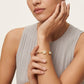 A close-up of a model wearing a delicate gold bangle bracelet with two round bezel-set stones on her wrist, paired with a light gray pleated sleeveless top.