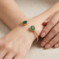 Two gold bangle bracelets with green stone inlays are shown on a model's wrist and hand against a neutral beige fabric background.