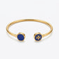 A gold-tone open cuff bangle bracelet with circular blue enamel end caps, one featuring a gold cross logo and the other a solid blue center, on a light gray background.