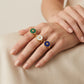 Three gold rings with round enamel inlays are shown on a person's hand. One ring has a green enamel background with a gold monogram, one has a white enamel background with a gold monogram, and one has a deep blue enamel background with a gold monogram.