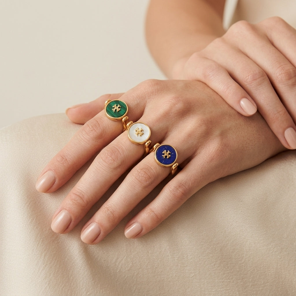 Three gold rings with round enamel inlays are shown on a person's hand. One ring has a green enamel background with a gold monogram, one has a white enamel background with a gold monogram, and one has a deep blue enamel background with a gold monogram.