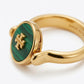 A gold ring featuring a round green malachite inlay with a gold monogram logo, shown on a clean white background.