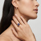 A close-up lifestyle shot of a model wearing a gold ring with a circular blue enamel center and a small gold star, alongside a matching gold huggie hoop earring.