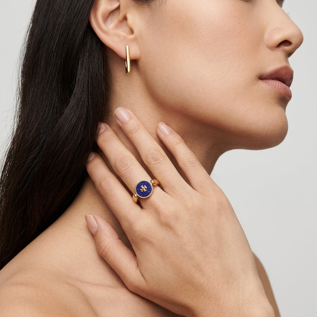 A close-up lifestyle shot of a model wearing a gold ring with a circular blue enamel center and a small gold star, alongside a matching gold huggie hoop earring.