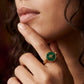 A close-up lifestyle shot of a model wearing a gold ring with a round green inlay featuring a gold monogram logo.