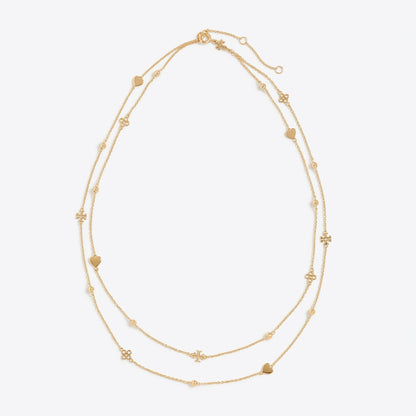 A delicate double-strand gold necklace featuring small heart and flower charms, displayed on a clean, light gray background.