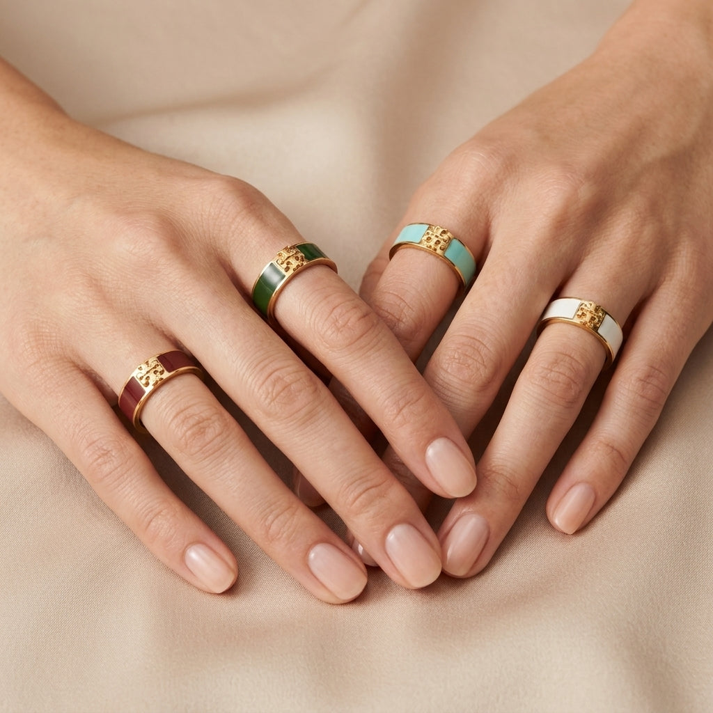Four rings displayed on a model's hands against a soft beige fabric background. The rings feature gold-toned metal bands with various colored inlays: one burgundy, one green, one light blue, and one white.