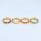 Four gold-plated rings with colorful enamel accents (black, orange, and turquoise) arranged in a row on a light gray background.