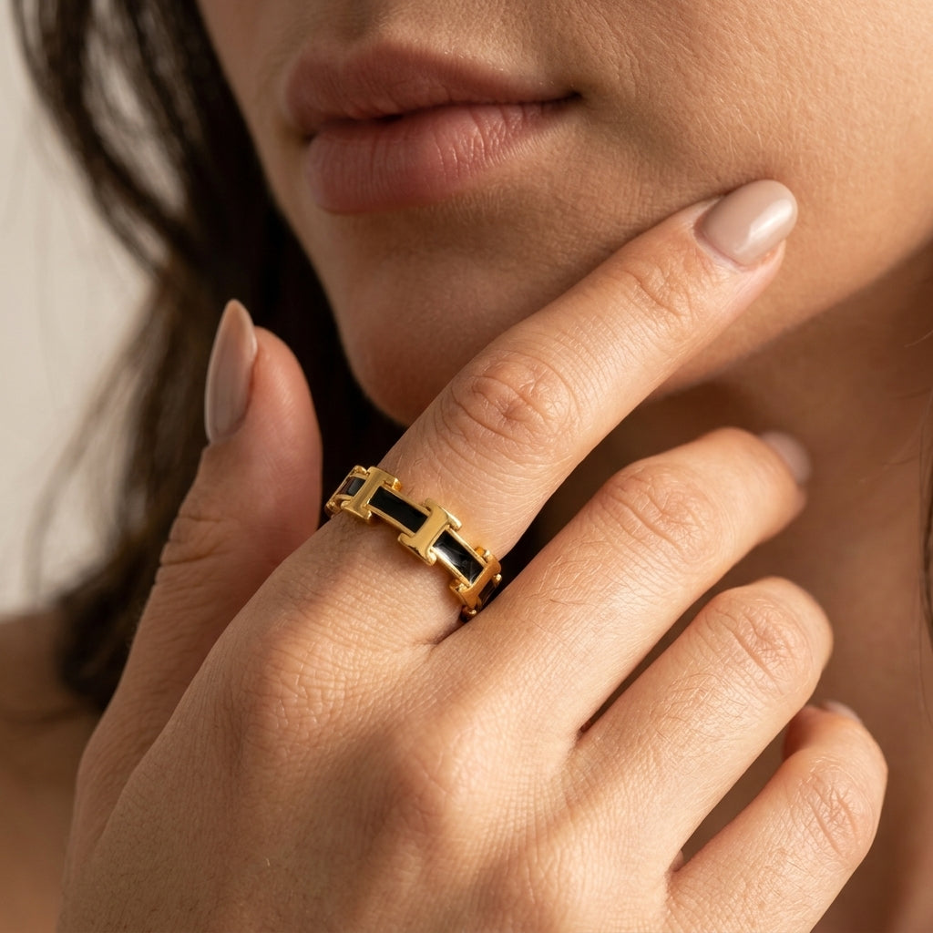 A close-up of a model's hand wearing a gold ring with black rectangular inlays, set against her face and neck.