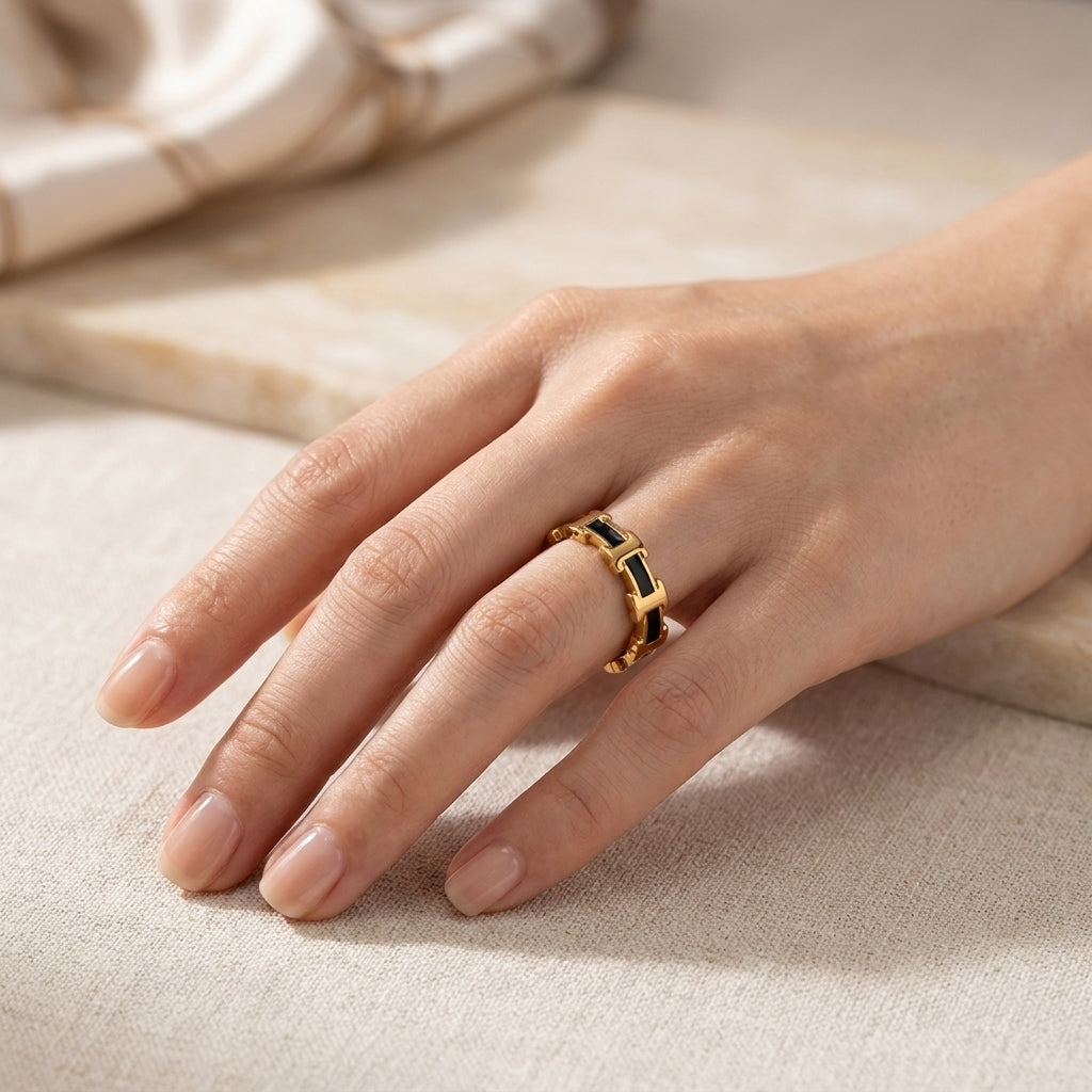 A gold ring with rectangular black inlay details, worn on a woman's hand resting on a neutral beige textured surface.