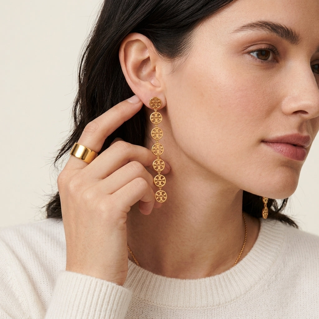 A close-up of a woman wearing a long gold drop earring with multiple circular logo-style motifs, along with a matching gold ring and necklace, against a soft light beige background.