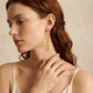 A close-up of a woman with red hair wearing a long, dangling gold earring with a repeating floral pattern, set against a soft beige background.