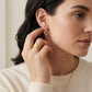 A close-up lifestyle shot of a woman wearing a delicate rose gold geometric dangle earring, with her hand touching her ear. She is wearing a cream-colored ribbed sweater and a gold ring.