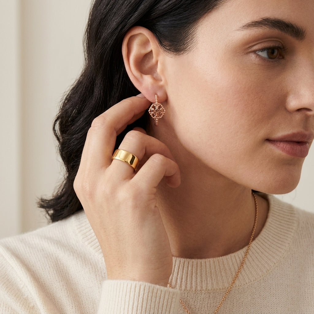 A close-up lifestyle shot of a woman wearing a delicate rose gold geometric dangle earring, with her hand touching her ear. She is wearing a cream-colored ribbed sweater and a gold ring.