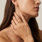 A close-up lifestyle shot of a model wearing a gold ring with a circular interlocking monogram design on her finger, alongside a matching gold bar earring.