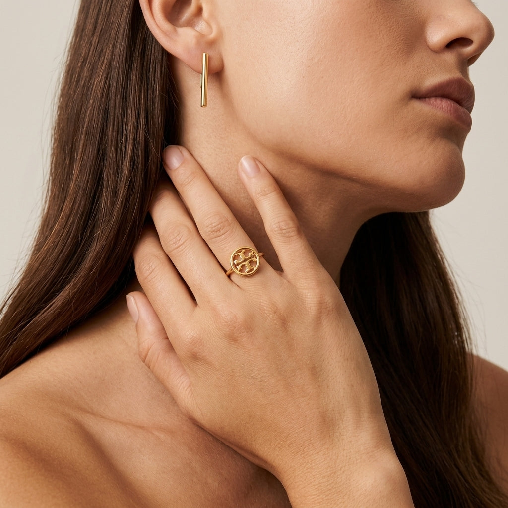 A close-up lifestyle shot of a model wearing a gold ring with a circular interlocking monogram design on her finger, alongside a matching gold bar earring.