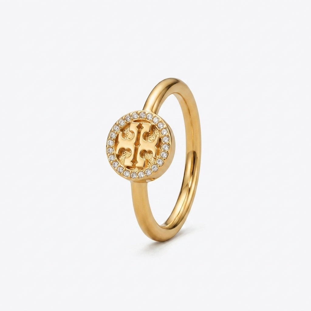 A gold ring featuring a circular emblem with a central stylized letter 'T' surrounded by a halo of small clear stones, set against a plain white background.