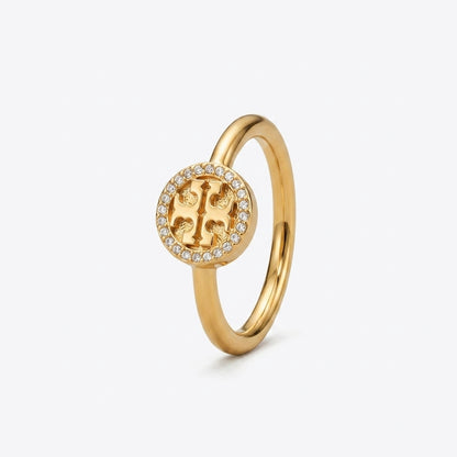 A gold ring featuring a circular emblem with a central stylized letter 'T' surrounded by a halo of small clear stones, set against a plain white background.