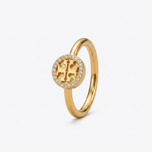 A gold ring featuring a circular emblem with a central stylized letter 'T' surrounded by a halo of small clear stones, set against a plain white background.