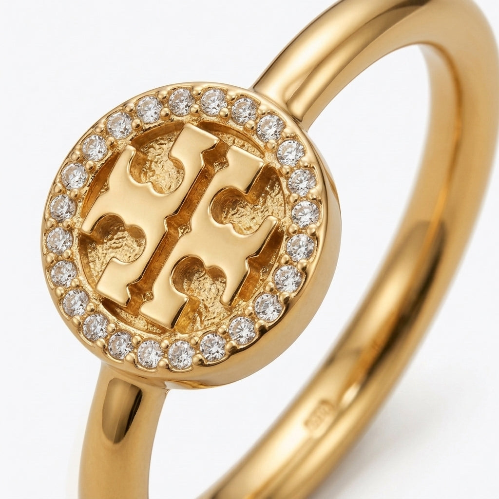 A gold ring featuring a circular face with a raised monogram logo surrounded by a border of small clear stones, shown on a clean white background.