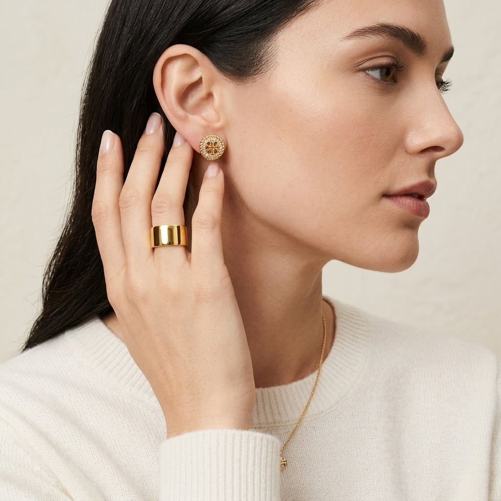 A close-up lifestyle shot of a woman wearing a gold ring and matching gold stud earring. The earring features a textured, floral-like gold pattern, while the ring is a wide, polished gold band.