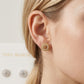 A close-up of a model's ear wearing a gold post stud earring with a circular logo medallion, with a blurred pair of matching earrings in the background.