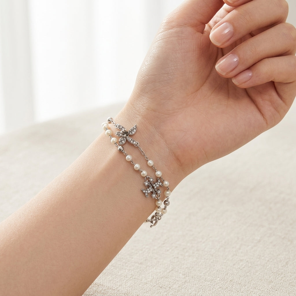 A delicate silver-toned double-strand bracelet with white pearls and crystal-embellished floral and cross motifs, shown worn on a woman's wrist.