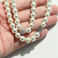 A close-up of a hand holding a double-strand pearl necklace with a silver-toned round pavé bead and a floral cluster charm.