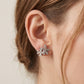 A close-up of a person's ear wearing a silver-toned stud earring with three small snowflake-like clusters set with clear stones, against a soft light beige background.